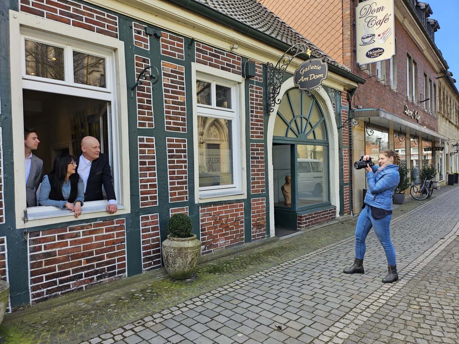 Eine Fotografin in blauer Jacke fotografiert drei Personen, die aus einem offenen Fenster eines historischen Backsteingebäudes mit dem Schild „Galerie Am Dom“ schauen. Die Szene spielt sich in einer gepflasterten Altstadtstraße neben einem Café und einer Kunstgalerie ab.