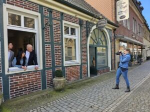 Die Fotografin macht ein Foto von drei Personen, die aus dem offenen Fenster eines historischen Backsteingebäudes schauen. Das Gebäude befindet sich an einer gepflasterten Gasse, und über dem Eingang ist ein Schild mit der Aufschrift „Dom Café“ zu sehen. Die Personen am Fenster lächeln und schauen zur Fotografin, die Atmosphäre ist locker und freundlich.
