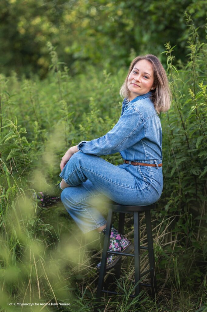 Eine Frau in einem Jeans-Overall sitzt seitlich auf einem schwarzen Hocker inmitten von grünem Pflanzenwuchs. Sie lächelt sanft und schaut in Richtung Kamera. Sie hat schulterlanges, helles Haar und bunte, geblümte Schuhe. Das Foto wurde in einer natürlichen Umgebung aufgenommen.