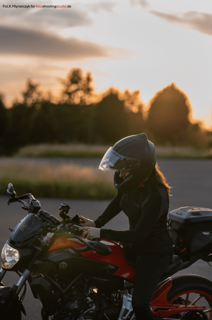 A woman in black motorcycle gear and helmet sits on a red and black Yamaha motorcycle during golden hour. The sun sets behind her, casting warm light and creating a serene, atmospheric scene. The background features soft-focus greenery.