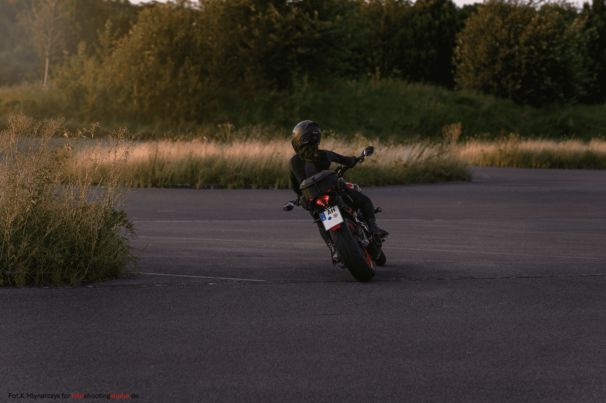 A motorcyclist in black riding gear and helmet is taking a curve on a practice lot. The photo is taken from behind in the warm light of sunset. Tall grass and a forested slope in the background create a natural, outdoor training atmosphere.