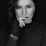 Black and white portrait of a woman with long dark hair, wearing a long-sleeved speckled top. She looks directly at the camera with a serious expression, her hand gently resting near her lips. She wears bracelets on one wrist and an earring is visible on one ear. The background is dark, highlighting her face and expression.