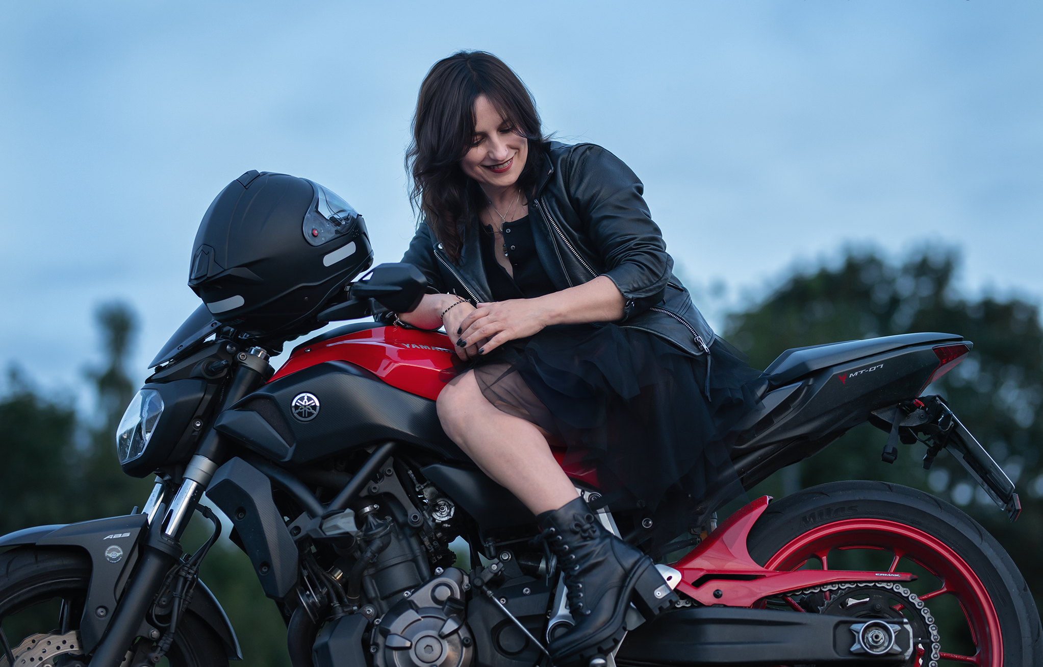 Eine Frau in schwarzer Lederjacke und Tüllkleid sitzt lächelnd auf einem rot-schwarzen Yamaha MT-07 Motorrad. Sie blickt nach unten und stützt sich mit der Hand auf den Tank. Im Hintergrund sind unscharfe Bäume und ein blauer Himmel zu sehen.