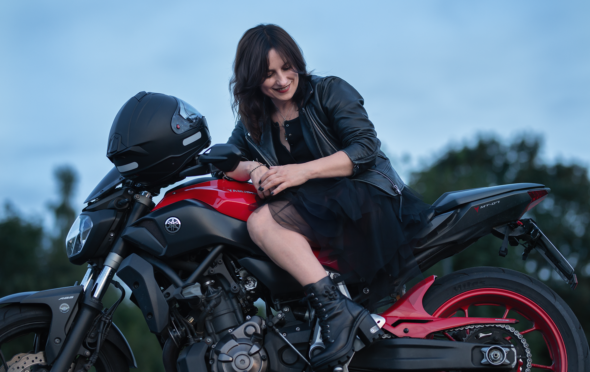 Kobieta w czarnej skórzanej kurtce i sukience siedzi na czerwono-czarnym motocyklu marki Yamaha MT-07. Uśmiecha się i patrzy w dół, trzymając rękę na zbiorniku paliwa. W tle widać rozmyte drzewa i niebieskie niebo.