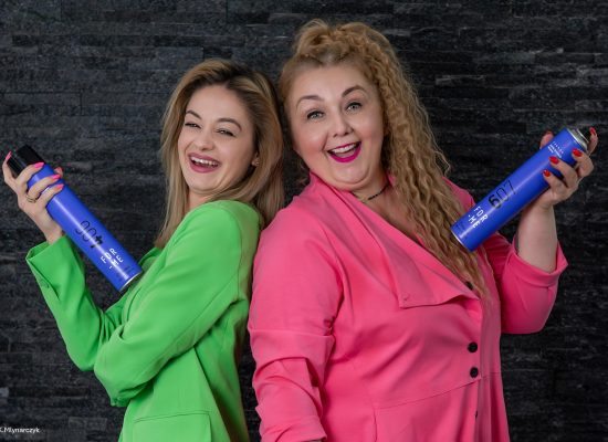 Business photography-hairdressing-salon-kameleon-products-hairspray-team.jpg Zwei lachende Frauen stehen Rücken an Rücken und halten blaue Haarspraydosen der Marke FOR ME in den Händen. Eine trägt einen grünen Anzug, die andere einen pinkfarbenen. Beide blicken fröhlich in die Kamera vor einem dunklen Hintergrund.