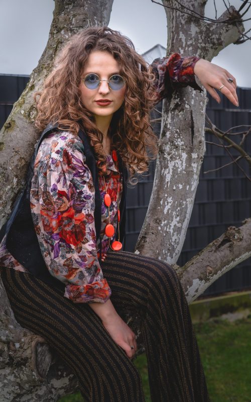 Eine junge Frau mit lockigen Haaren und runden Brillen sitzt auf einem Ast. Sie trägt eine gemusterte Bluse mit Blumenmotiv, eine dunkle Weste und gestreifte Hosen. Ihr Stil erinnert an Retro- oder Boho-Mode. Im Hintergrund ist ein dunkler Zaun und ein Stück Garten zu sehen.