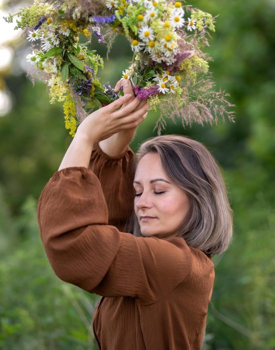 Porträtfotografie-authentisch-Leidenschaft-Ewa-Roman-Business-Natur-Wildblumenkranz-Frau-Aroma-Roh-Natur.jpg Eine Frau in einem braunen Kleid hält einen Kranz aus Wildblumen und Gräsern über ihren Kopf. Sie hat die Augen geschlossen und wirkt ruhig und entspannt. Im Hintergrund ist üppiges Grün zu sehen, was dem Bild eine natürliche, verträumte Atmosphäre verleiht.