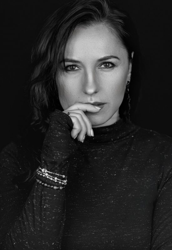 Black and white portrait of a woman with long dark hair, wearing a long-sleeved speckled top. She looks directly at the camera with a serious expression, her hand gently resting near her lips. She wears bracelets on one wrist and an earring is visible on one ear. The background is dark, highlighting her face and expression.