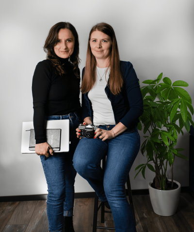 Zwei Frauen in lässiger Kleidung posieren in einem Fotostudio. Eine hält eine Kamera in der Hand, die andere ein Grafiktablet und ein Skizzenbuch. Beide lächeln dezent in die Kamera. Im Hintergrund ist eine Zimmerpflanze und eine weiße Wand zu sehen.
