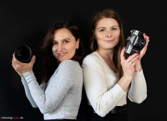 Zwei Frauen stehen Rücken an Rücken vor einem schwarzen Hintergrund und lächeln. Die Frau auf der linken Seite trägt ein graues Oberteil und hält ein Kameraobjektiv in Richtung des Betrachters. Die Frau auf der rechten Seite trägt einen cremefarbenen Pullover und hält eine klassische analoge Kamera vom Typ Zenit-E. Beide wirken professionell und selbstbewusst – ein eingespieltes kreatives Fotografen-Team.
