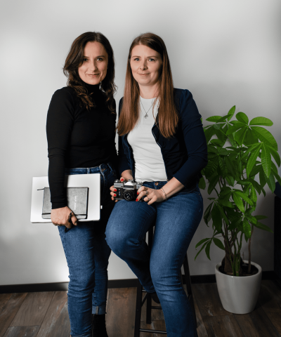 Two women in casual clothing pose in a photo studio. One is holding a camera, the other a graphic tablet and sketchbook. Both smile gently at the camera. A potted plant and a white wall are visible in the background.