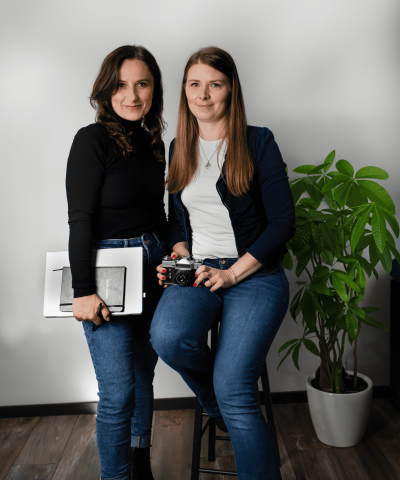 Two women in casual clothing pose in a photo studio. One is holding a camera, the other a graphic tablet and sketchbook. Both smile gently at the camera. A potted plant and a white wall are visible in the background.