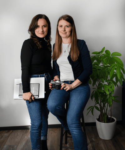 Dwie kobiety w codziennych ubraniach pozują w studiu fotograficznym. Jedna z nich trzyma aparat fotograficzny, druga – tablet graficzny i szkicownik. Obie uśmiechają się subtelnie do obiektywu. W tle widoczna jest roślina doniczkowa i biała ściana.