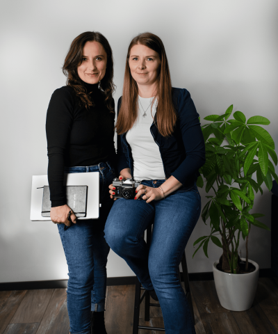Dwie kobiety w codziennych ubraniach pozują w studiu fotograficznym. Jedna z nich trzyma aparat fotograficzny, druga – tablet graficzny i szkicownik. Obie uśmiechają się subtelnie do obiektywu. W tle widoczna jest roślina doniczkowa i biała ściana.