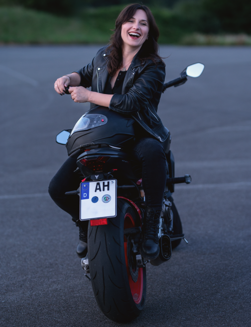 hobby-fotografia-andzelika.sawka-kobieta-motocykl-śmiejąca się-skórzana kurtka-sesja-zdjęciowa-outdoor.jpg Młoda kobieta w czarnej skórzanej kurtce siedzi tyłem na motocyklu sportowym, uśmiechając się szeroko i patrząc w stronę aparatu. Ma ciemne włosy i emanuje radością oraz pewnością siebie. Zdjęcie wykonane na pustym placu asfaltowym o zmierzchu.