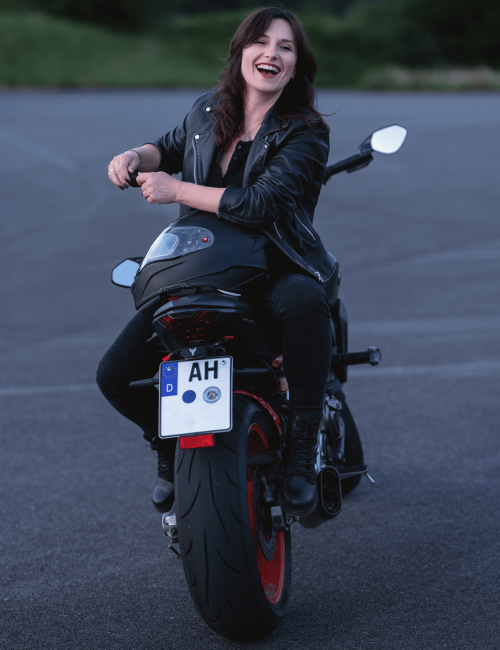 hobby-photography-andzelika.sawka-woman-motorcycle-laughing-leather-jacket-photoshoot-outdoor.jpg A young woman in a black leather jacket sits facing backward on a sport motorcycle, smiling brightly and looking at the camera. She has dark hair and radiates joy and confidence. The photo was taken at dusk on an empty asphalt lot.
