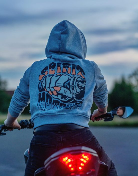 photography-hobby-andzelika-sawka-motorcyclist-spirit-hoody-sunset.jpg A person in a grey hoodie sits on a motorcycle facing away, ready to ride. The back of the hoodie features a colorful graphic of a clenched fist gripping a throttle with the words “Serial Throttles.” The motorcycle’s red tail light glows brightly. The photo is taken at dusk.