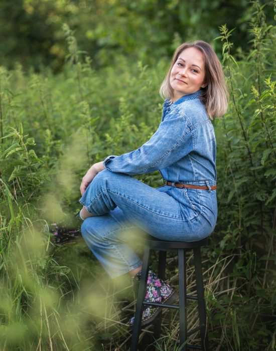 portrait-photography-authentic-passion-ewa-roman-business-nature-in a meadow of nettles-woman-aroma-raw-nature.jpg A woman in a denim jumpsuit sits sideways on a black stool surrounded by greenery. She smiles gently, looking toward the camera. She has shoulder-length light hair and colorful floral shoes. The photo was taken in a natural outdoor setting among tall plants.