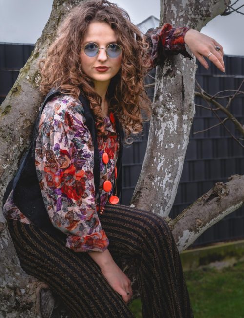 portrait-photography-nikola-czerniecka-boho-fashion-woman-tree-hippie-look-glasses-patterned-blouse.jpg A young woman with curly hair and round glasses sits on a tree branch. She is wearing a floral patterned blouse, a dark vest, and striped pants. Her style reflects a retro or boho fashion. A dark fence and part of a garden can be seen in the background.