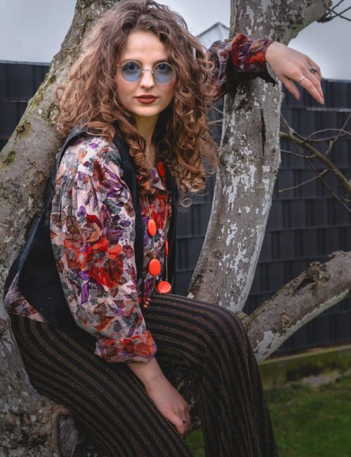 A young woman with curly hair and round glasses sits on a tree branch. She is wearing a floral patterned blouse, a dark vest, and striped pants. Her style reflects a retro or boho fashion. A dark fence and part of a garden can be seen in the background.