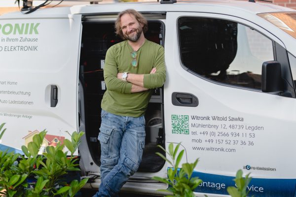 witronik-business-photography-witronik-witold-sawka-electrician-electrical engineer-smart-home-installation-e-car-charging-station.jpg A bearded man with long hair, wearing a green shirt and jeans, leans casually against a white company van with the door open, smiling at the camera. The van displays contact details for "Witronik", a company specializing in electronics and smart home solutions. Plants are visible in the foreground.